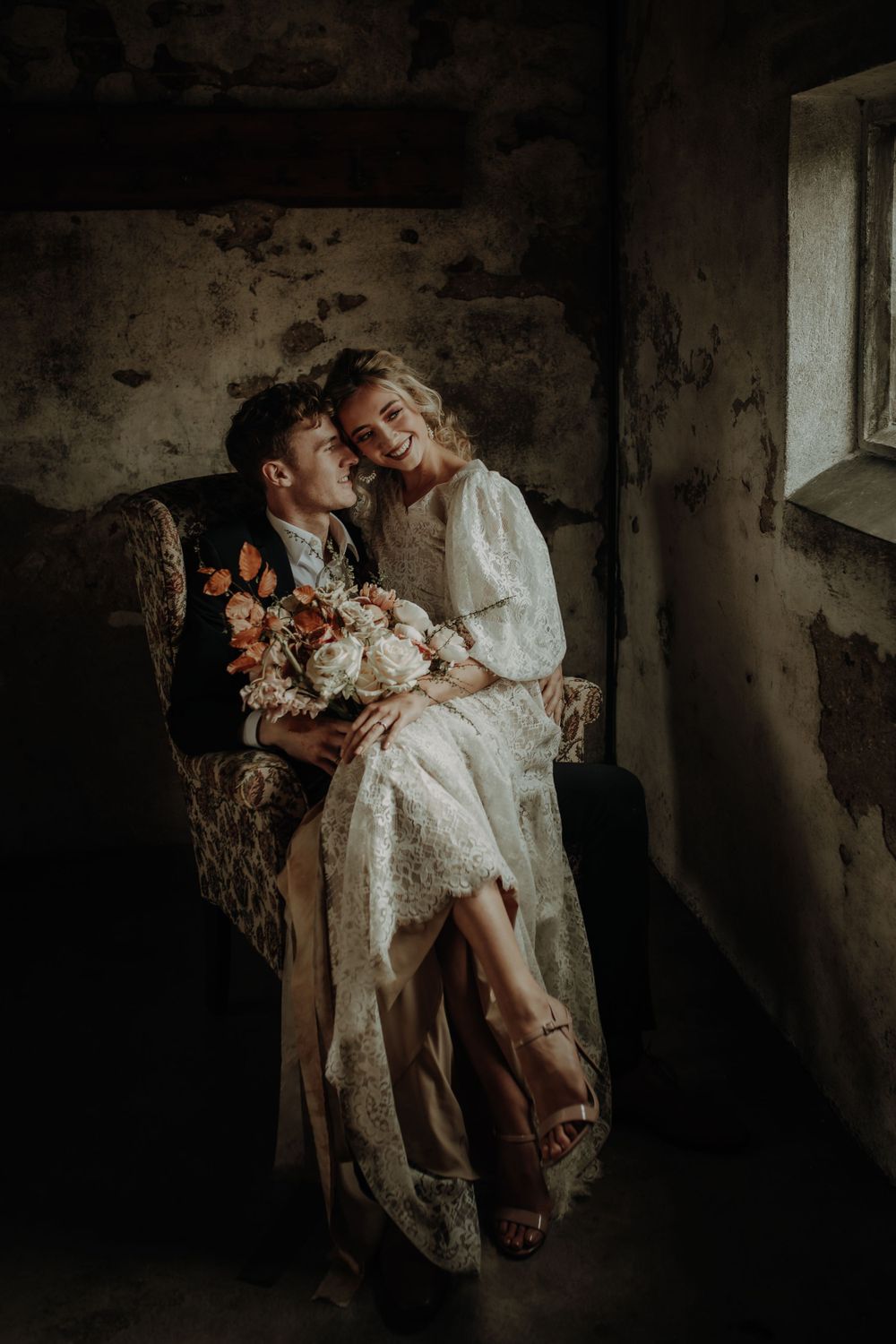 A romantic vintage-styled couple shares an intimate moment in a weathered armchair holding a floral bouquet.