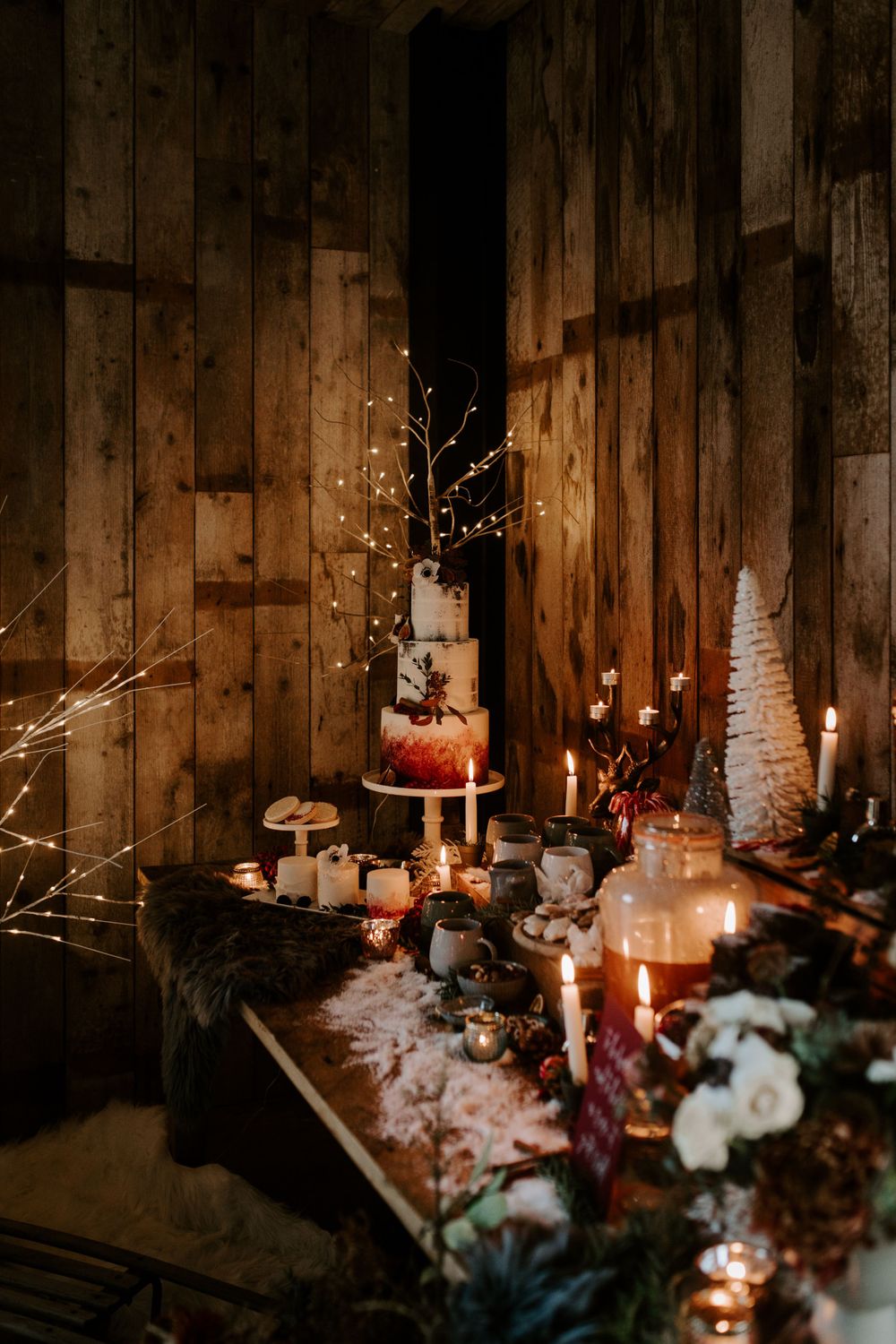 A rustic wedding dessert table with candles, fairy lights and a tiered cake against wooden walls.