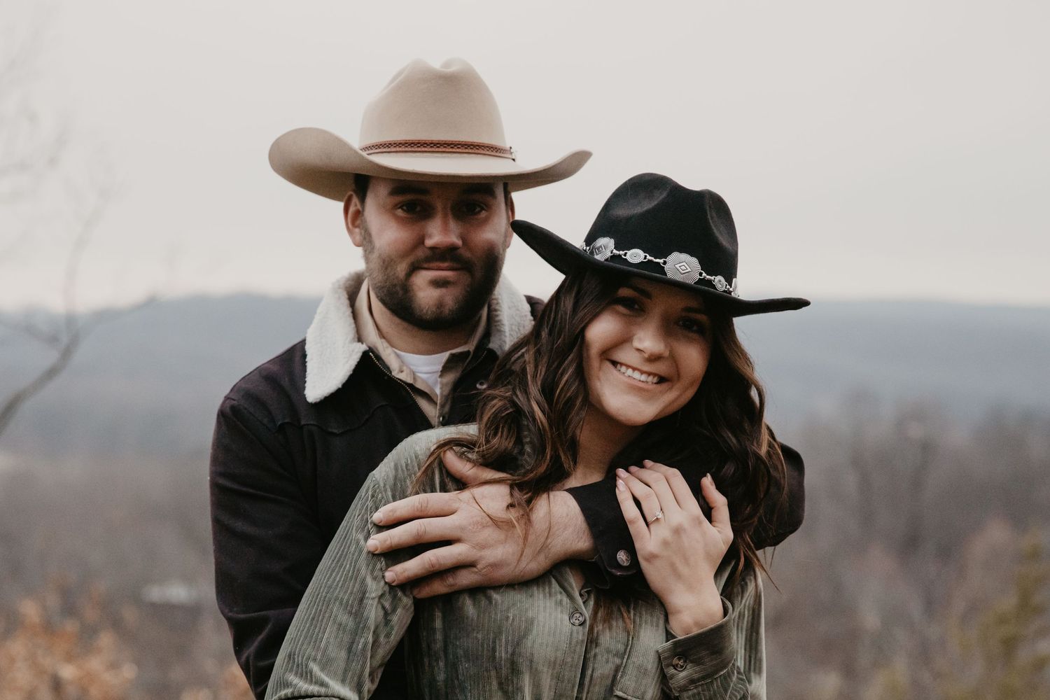 couple embraces each other and smiles at the camera, each wearing a trendy cowboy had and earth tone shirts, she wears her engagement ring