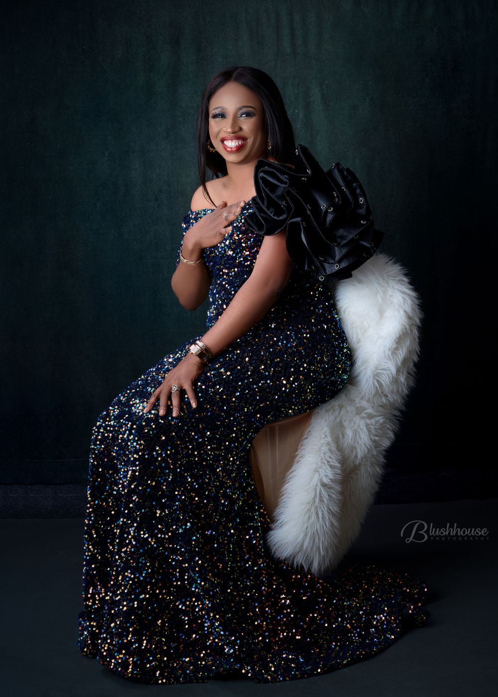 Elegant woman in sparkly navy gown poses on white fur chair against dark backdrop.