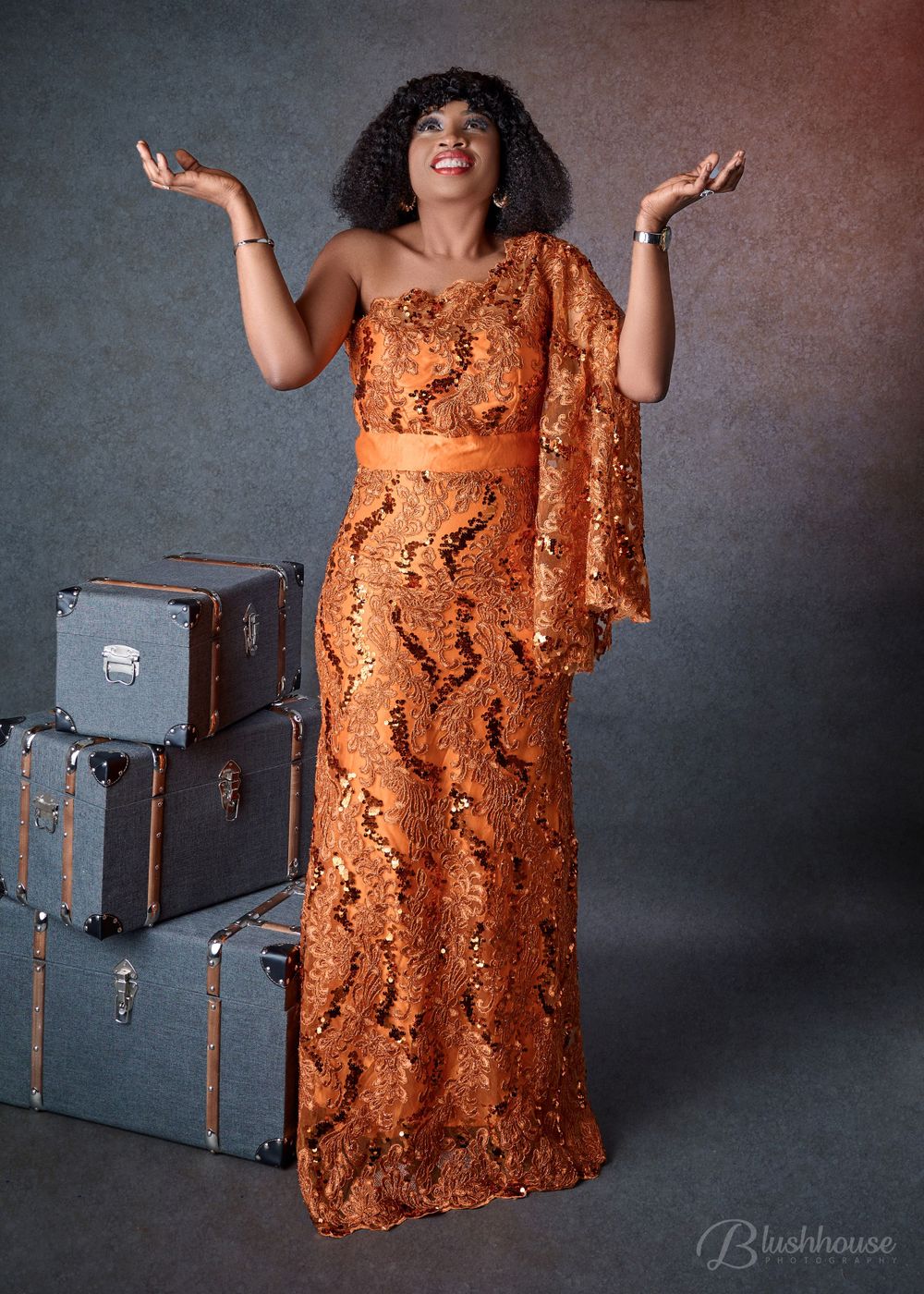 Woman in orange lace dress poses joyfully next to vintage suitcases.