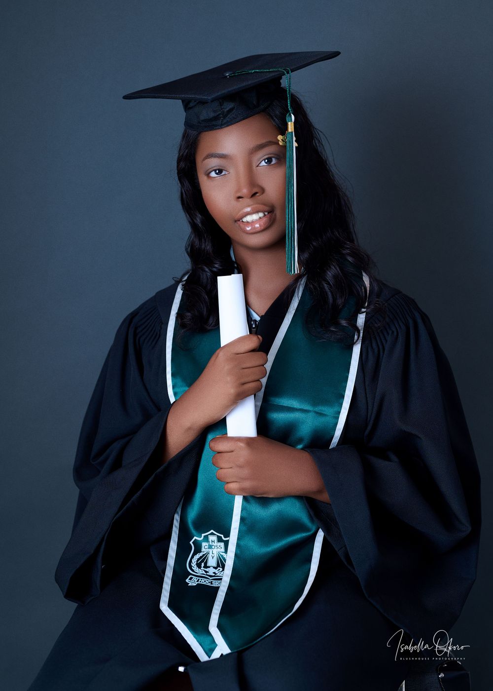Graduate in black cap and gown with emerald green sash holds diploma during professional portrait session.