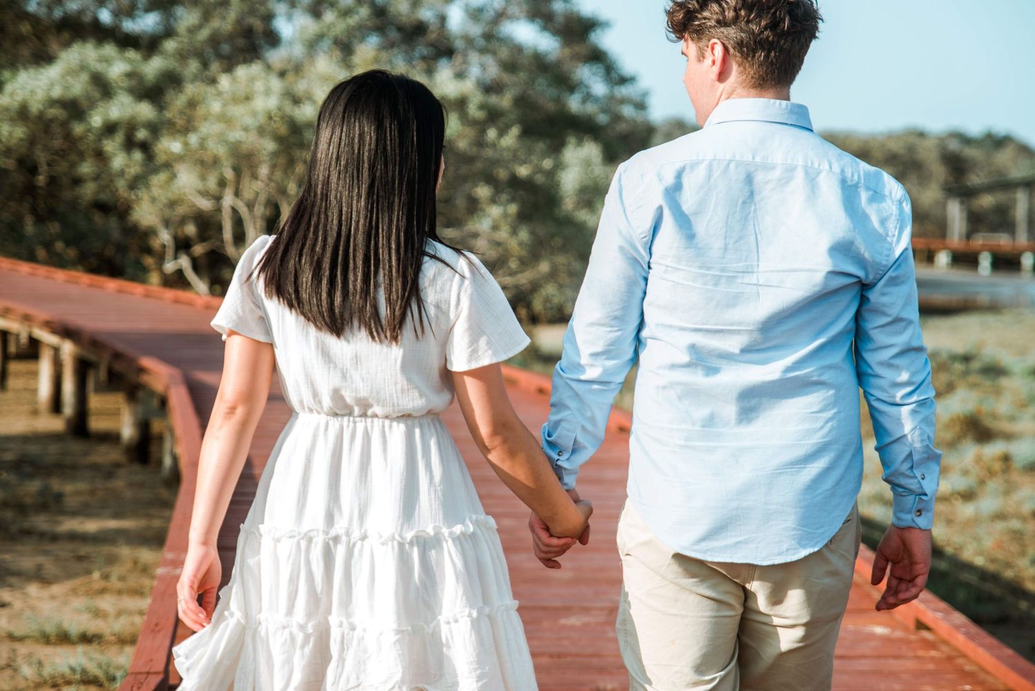 A couple walks hand in hand on a wooden boardwalk through coastal scenery.