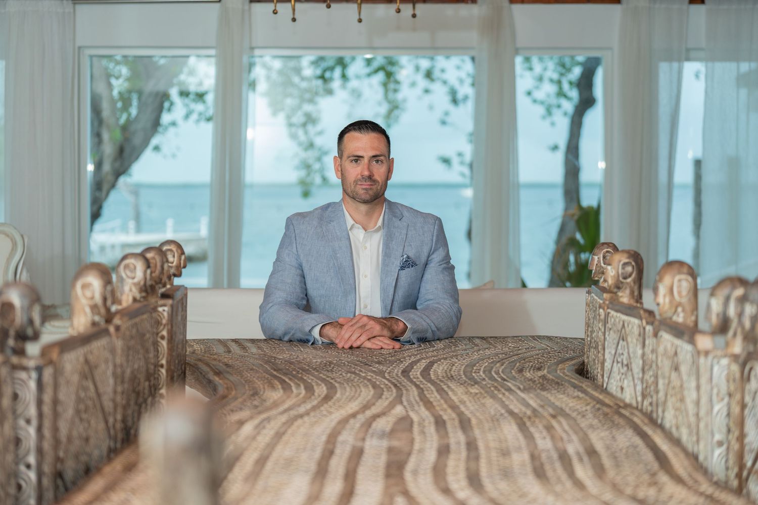 Professional portrait of someone in a gray suit seated at a wooden dining table with ornate chairs.