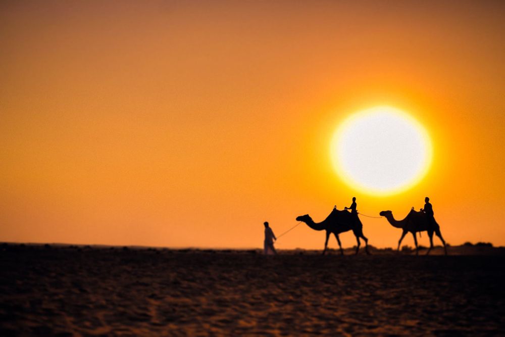 Silhouette of camels and riders crossing a vast desert landscape against a vibrant orange sunset.
