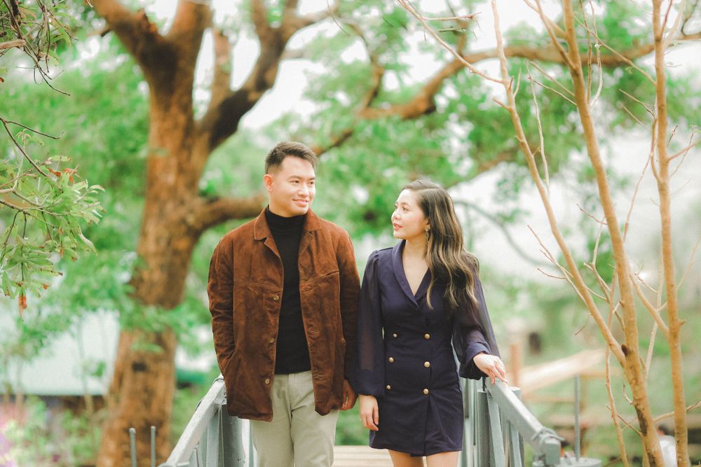 Stylish Asian couple walking on bridge surrounded by lush green trees in casual elegant outfits.