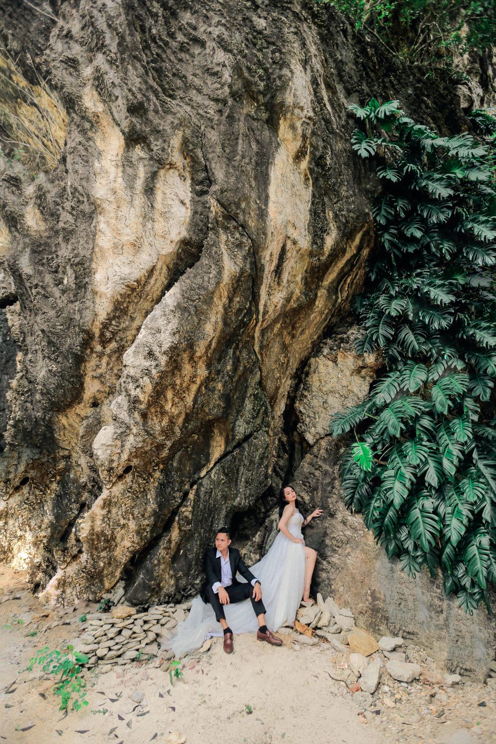 A bride and groom pose dramatically against a massive rocky cliff face surrounded by tropical greenery.