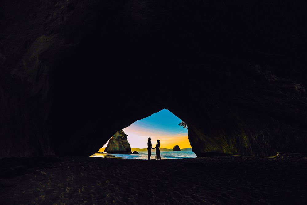Two silhouetted figures hold hands inside a dark cave with a stunning colorful sunset visible through the opening.