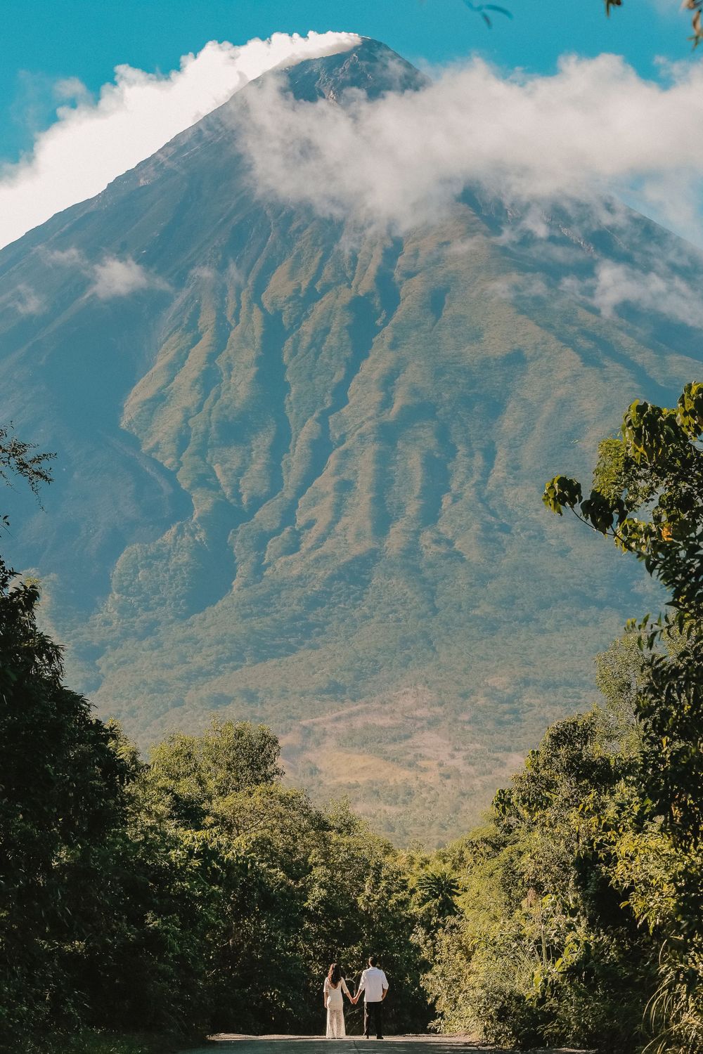 Couple holding hands before a majestic volcanic mountain landscape with lush green trees.