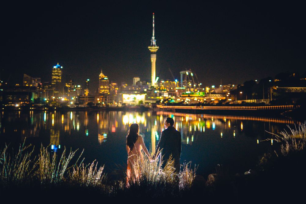 Couple standing by a reflective waterfront with a glowing city skyline at night.