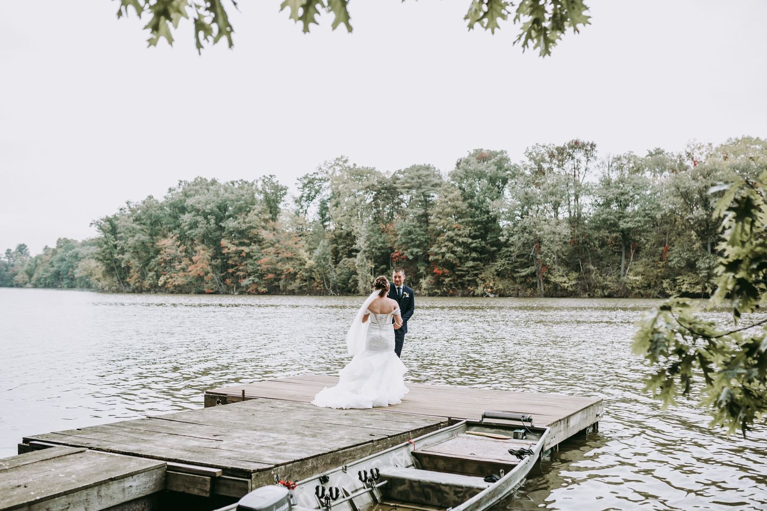 Wedding couple embracing on rustic dock with lake and forest backdrop.