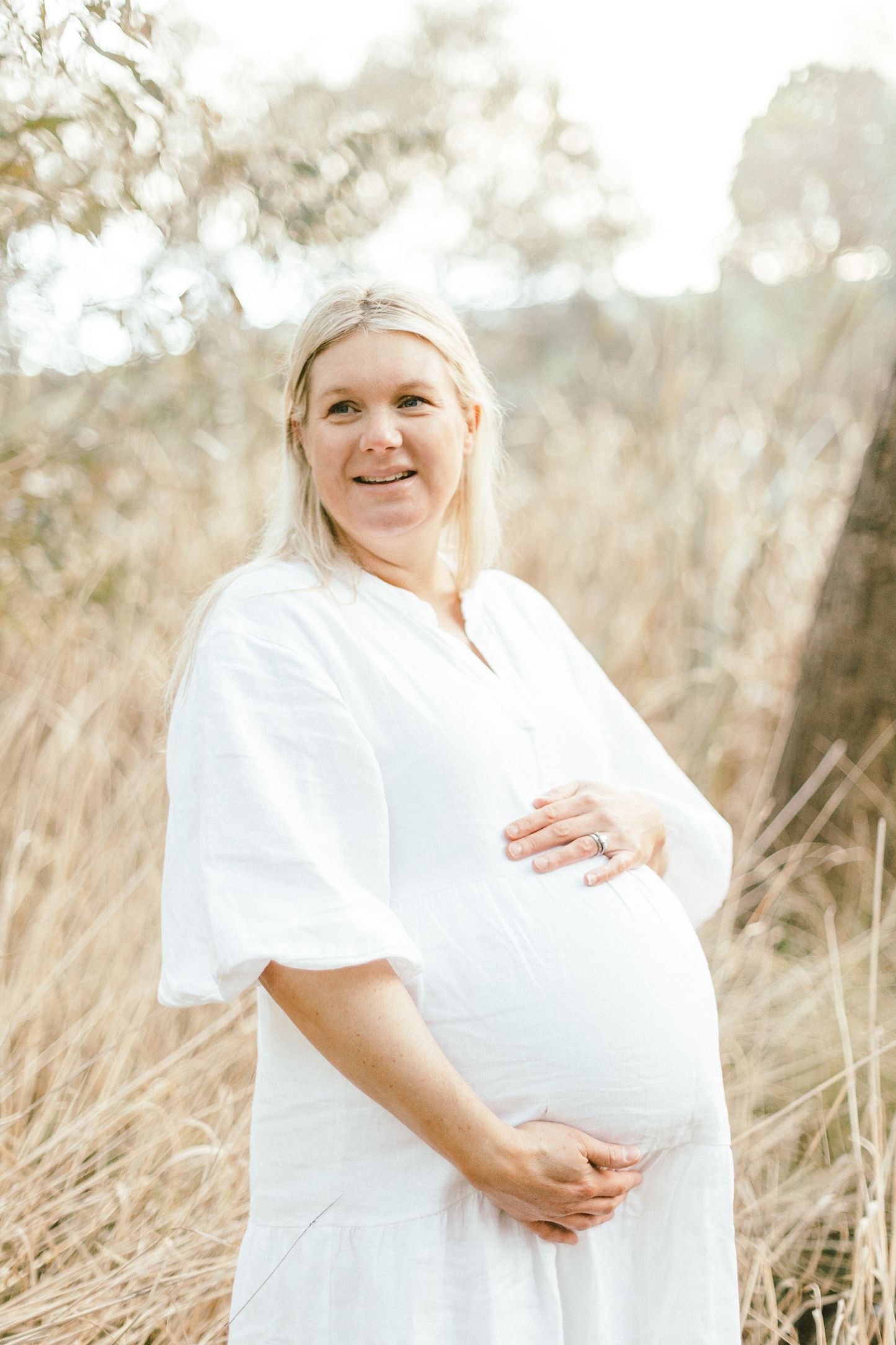 Radiant maternity portrait of woman in white dress surrounded by natural light.