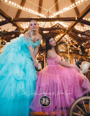 Two women in pastel blue and pink tulle gowns pose on a carousel at night.
