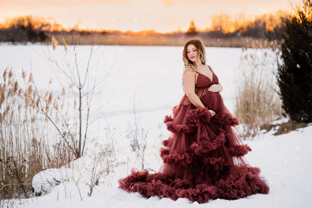 Maternity portrait featuring burgundy ruffled gown against snowy winter backdrop.