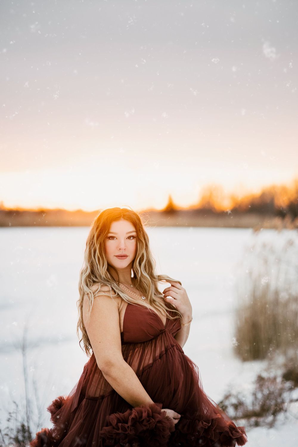 Winter maternity photo of woman in burgundy dress against snowy landscape at sunset.