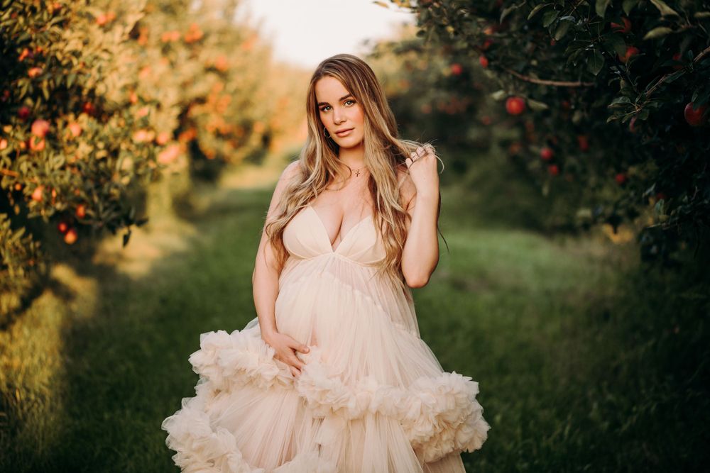 A pregnant woman in a blush pink tulle gown poses gracefully in an orchard during golden hour.