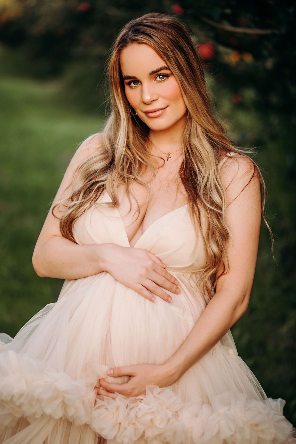 Expectant mother wearing a soft pink tulle gown poses in a garden setting.