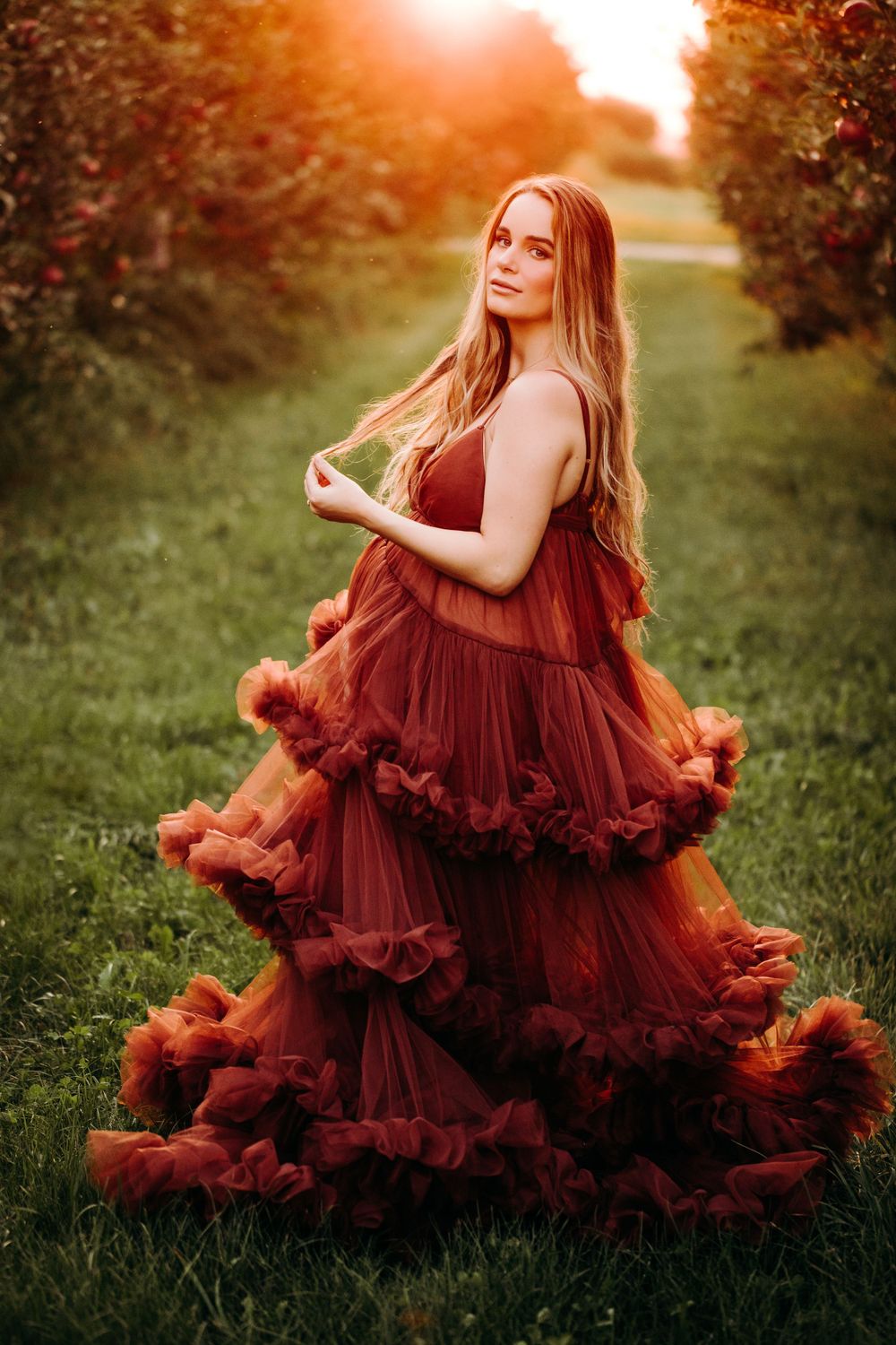 Woman in flowing burgundy gown poses between rows of trees during sunset.