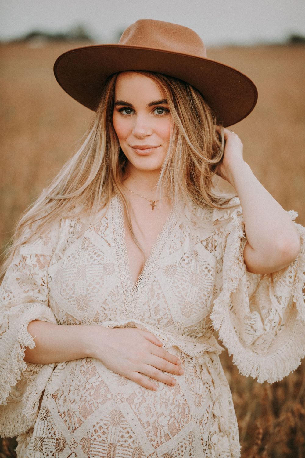 A woman in a lace dress and brown hat poses in a golden wheat field at sunset.