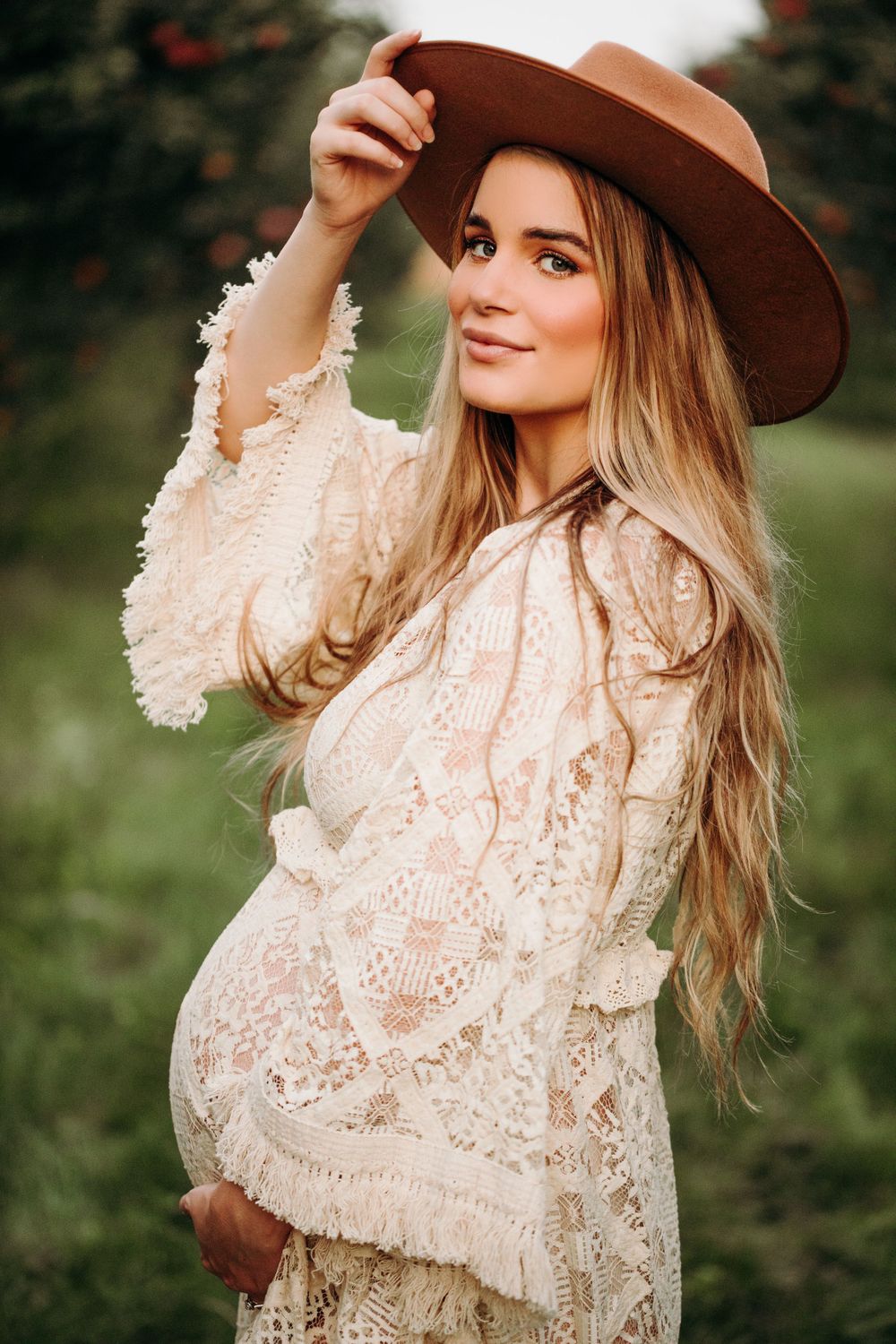 A maternity portrait with woman in cream lace dress and brown hat in a garden setting.