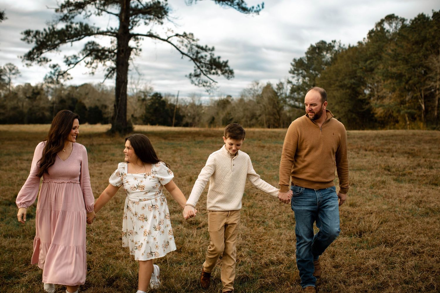 Family walking together hand in hand through an autumn field with pine trees in the background.