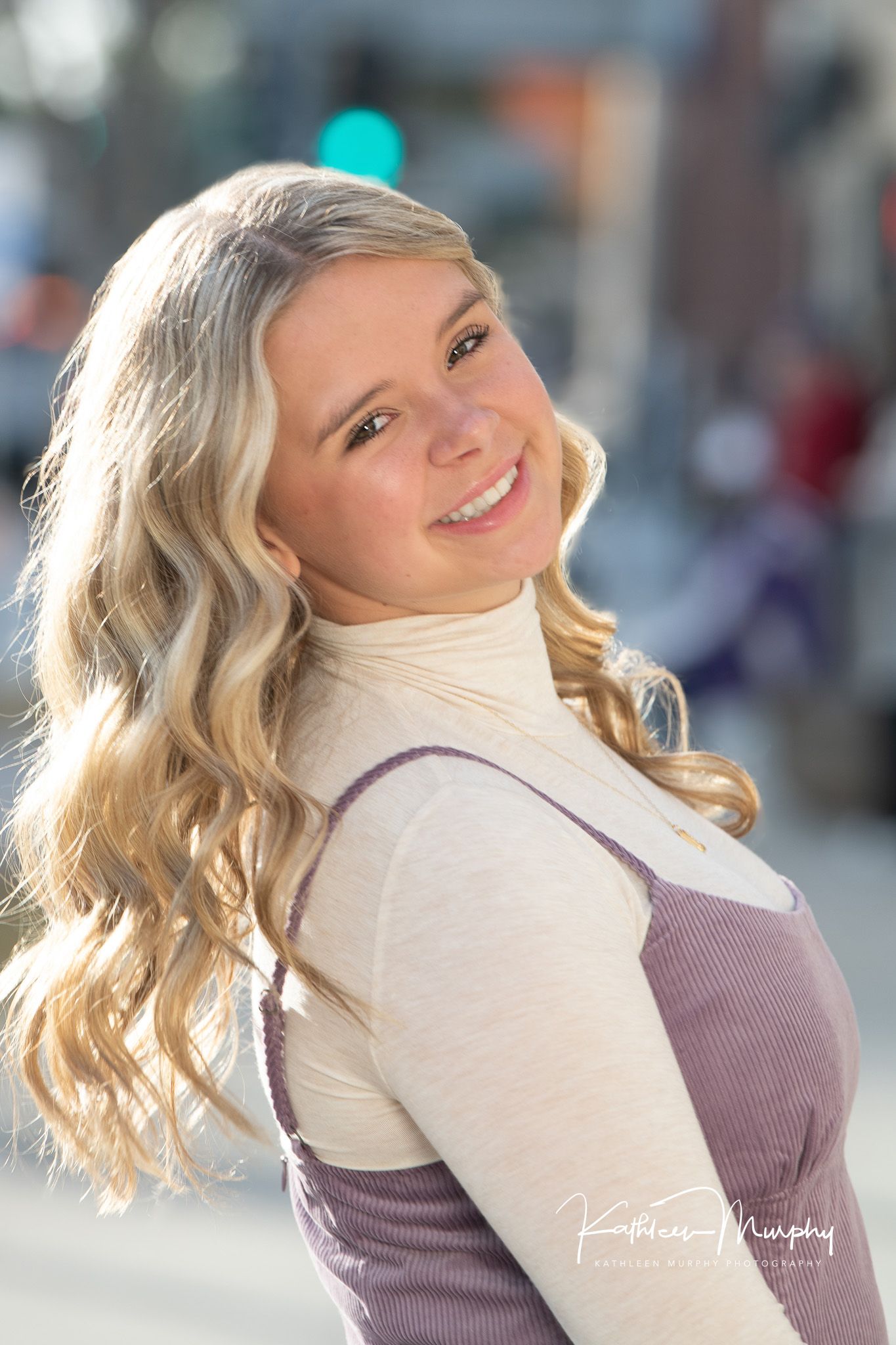 Close-up portrait showing wavy blonde hair and cream turtleneck with purple dress against blurred urban background.