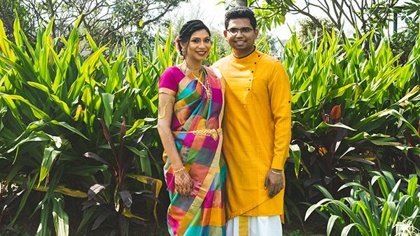 Couple in traditional Indian wedding attire standing on white balcony with flowering plants.