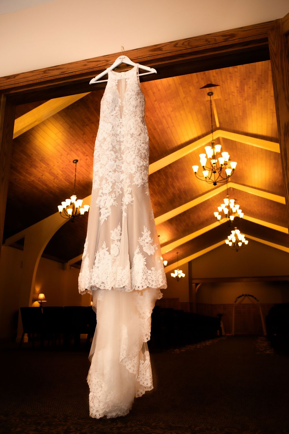 Lace wedding dress hangs in venue with warm lighting and vaulted wooden ceiling.