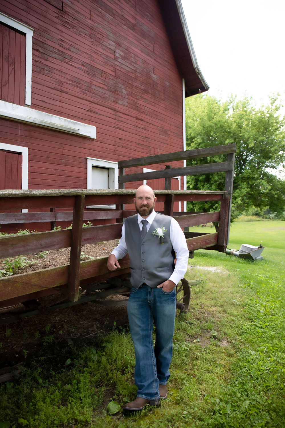 Groomsman in grey vest poses casually by red barn deck.