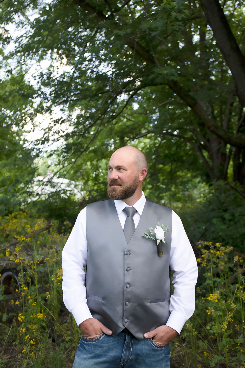 Groomsman in grey vest stands among wildflowers and trees.