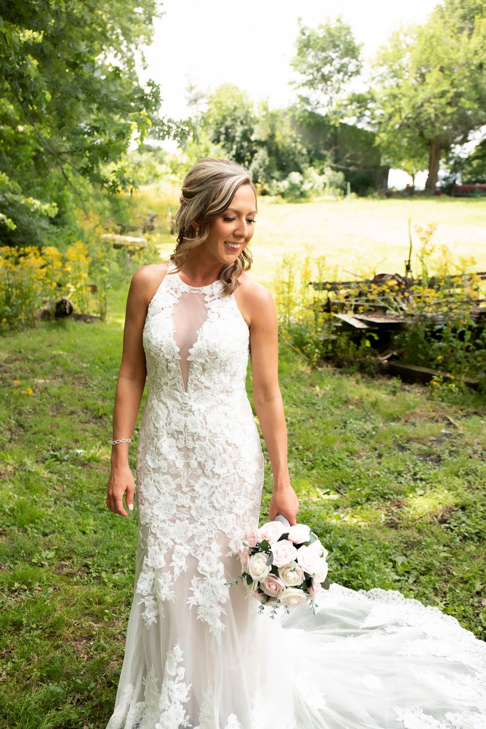 Elegant bride in lace wedding dress with floral appliques holds white bouquet in outdoor garden setting.