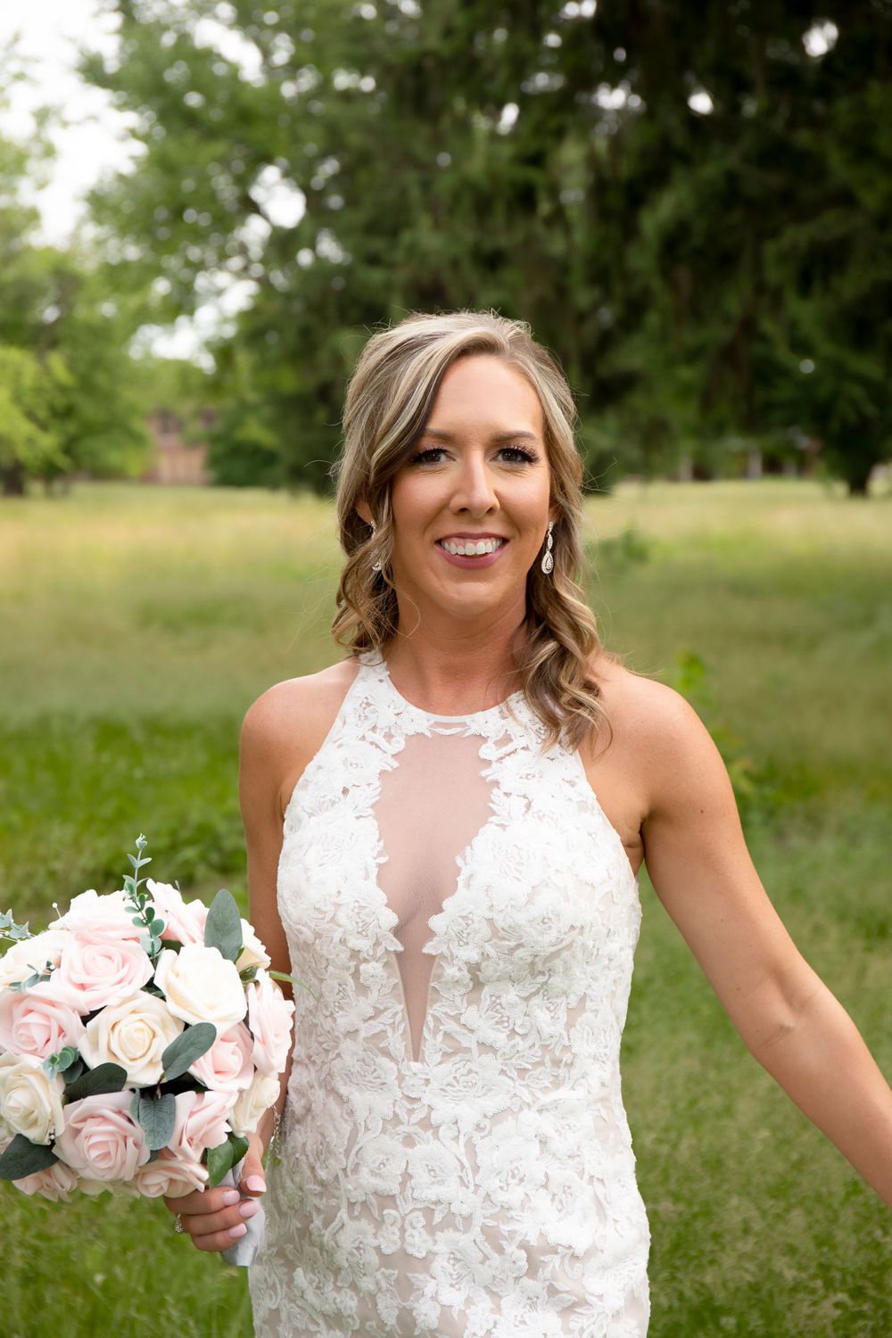 Bride in fitted lace halter wedding gown holds pink and white bouquet against garden backdrop.