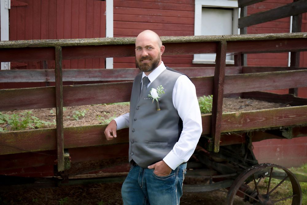 Groom in grey vest and white shirt stands near red barn in rustic setting.