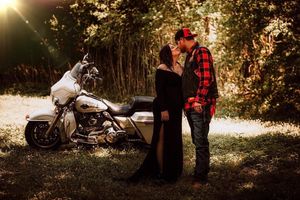 A romantic couple poses together next to a motorcycle on a sunlit forest path.