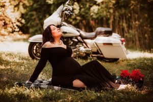 A woman in a black dress poses elegantly next to a white motorcycle in a sunlit outdoor setting.