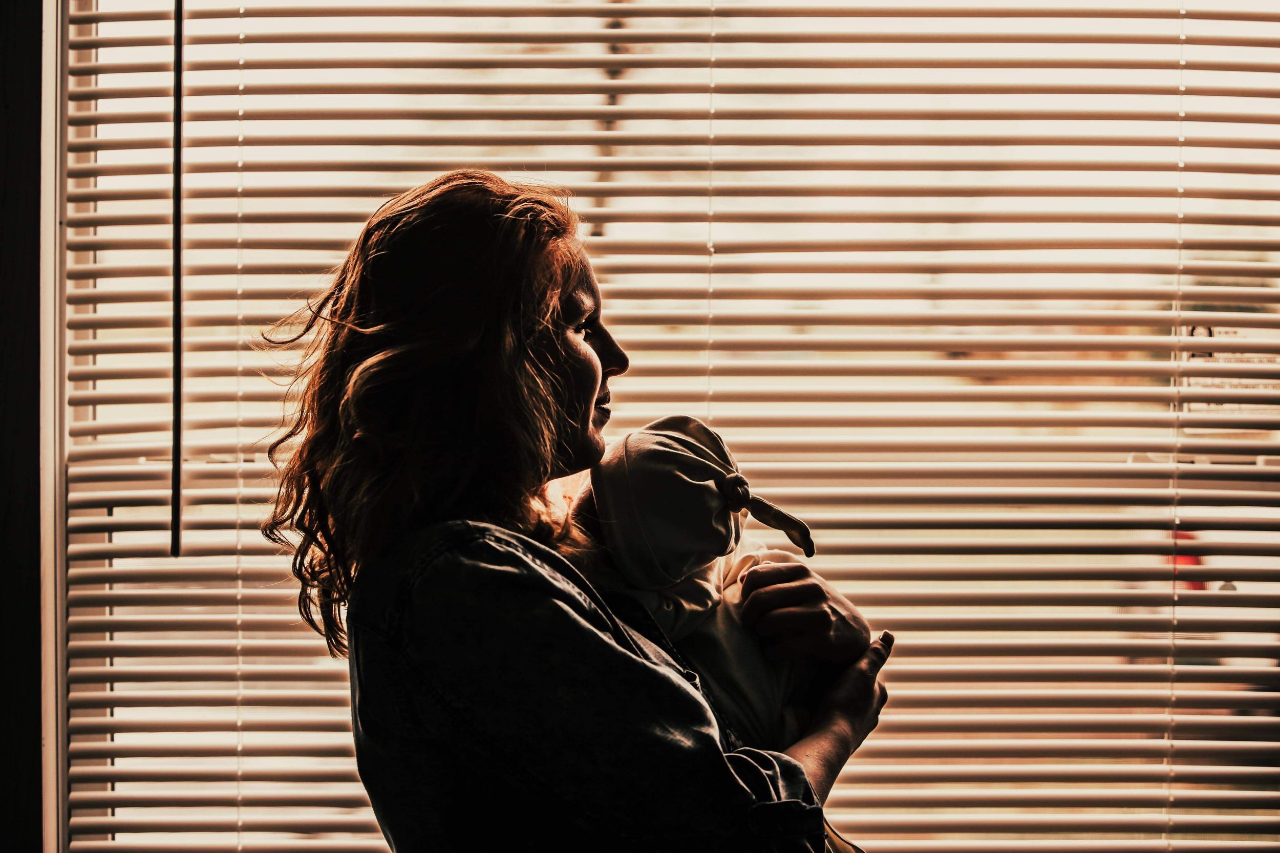 A silhouetted figure holds a baby against window blinds in warm lighting.