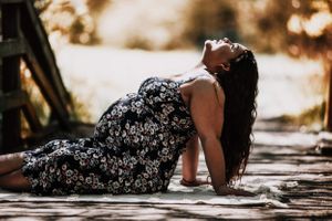 A artistic maternity photo of a woman in a floral dress reclining on wooden planks in sunlight.