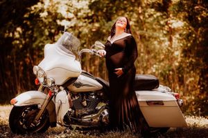 A maternity photoshoot featuring a woman in a black dress posing with a white motorcycle outdoors.