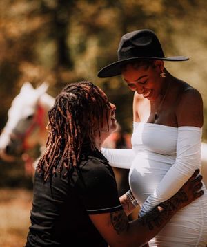A tender maternity moment captured outdoors with a white dress and black hat against an autumn background.