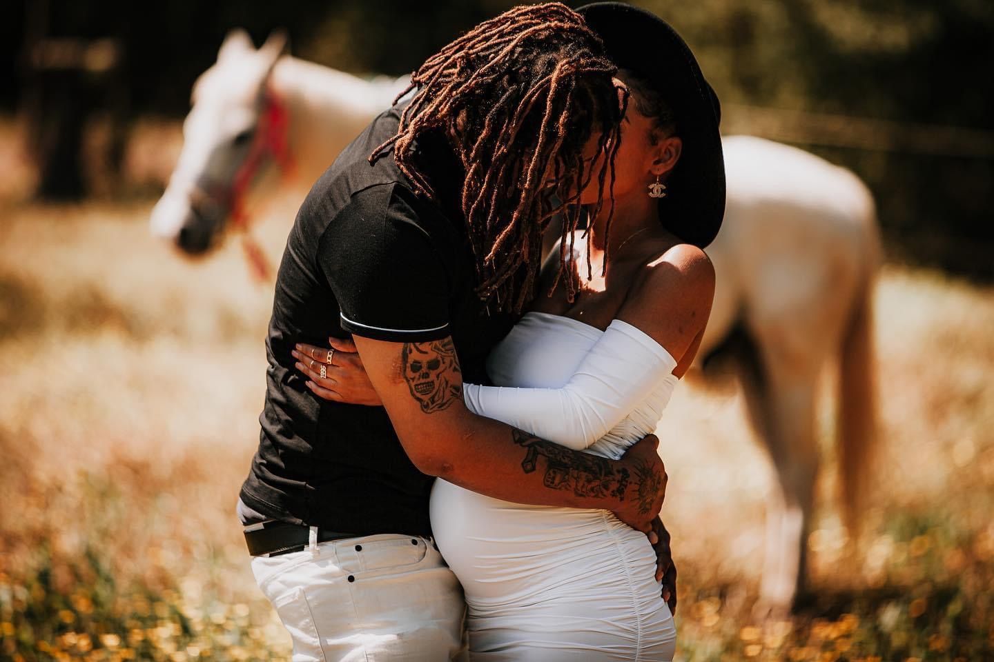 An intimate embrace captured outdoors with a white horse visible in the background.