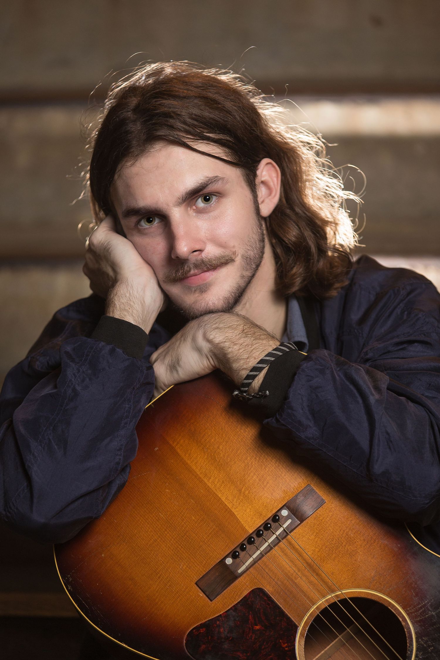 Musician poses with acoustic guitar in sunburst finish against rustic wooden background.