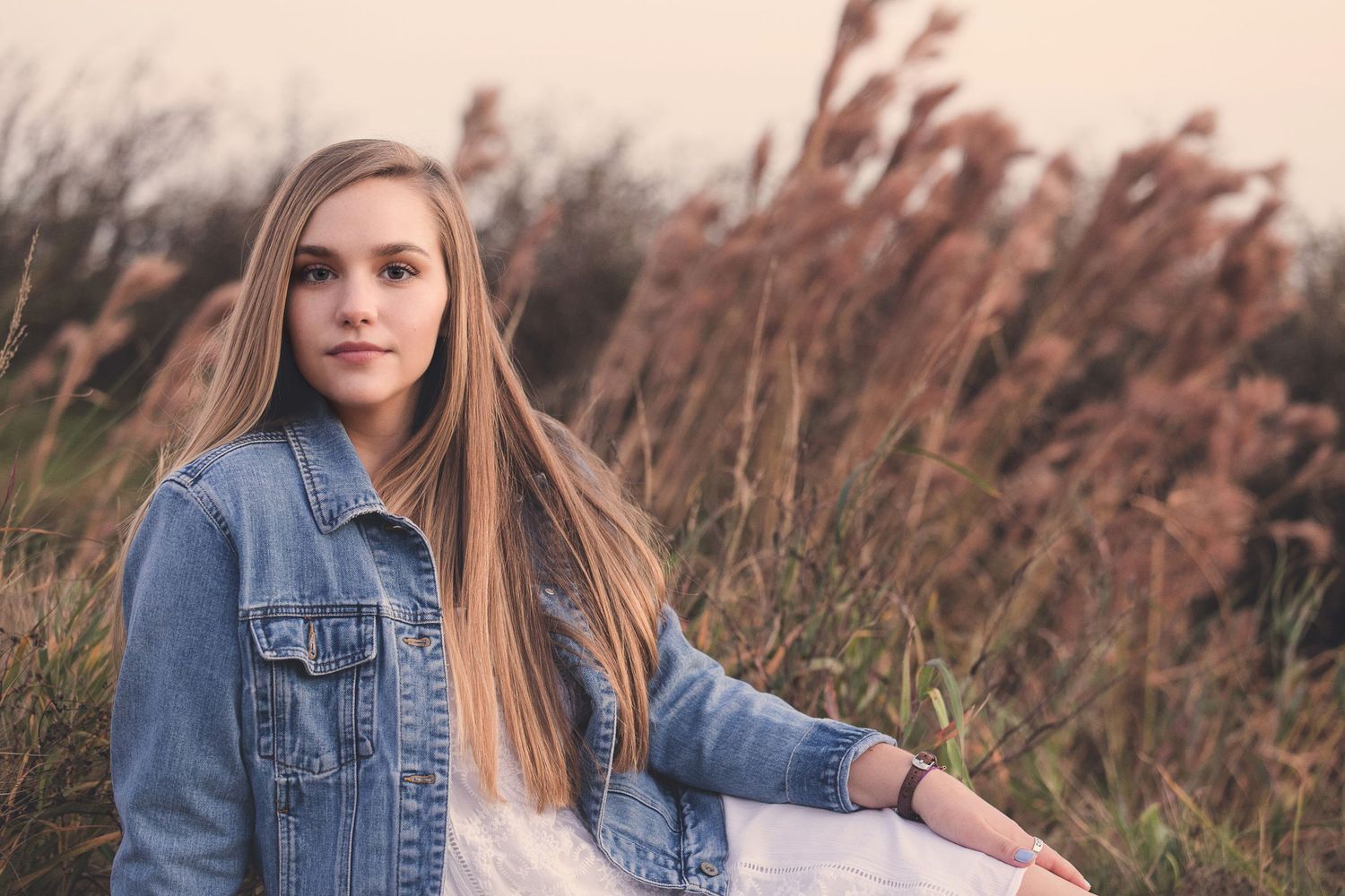 Young woman in denim jacket poses in field of tall grass during golden hour.