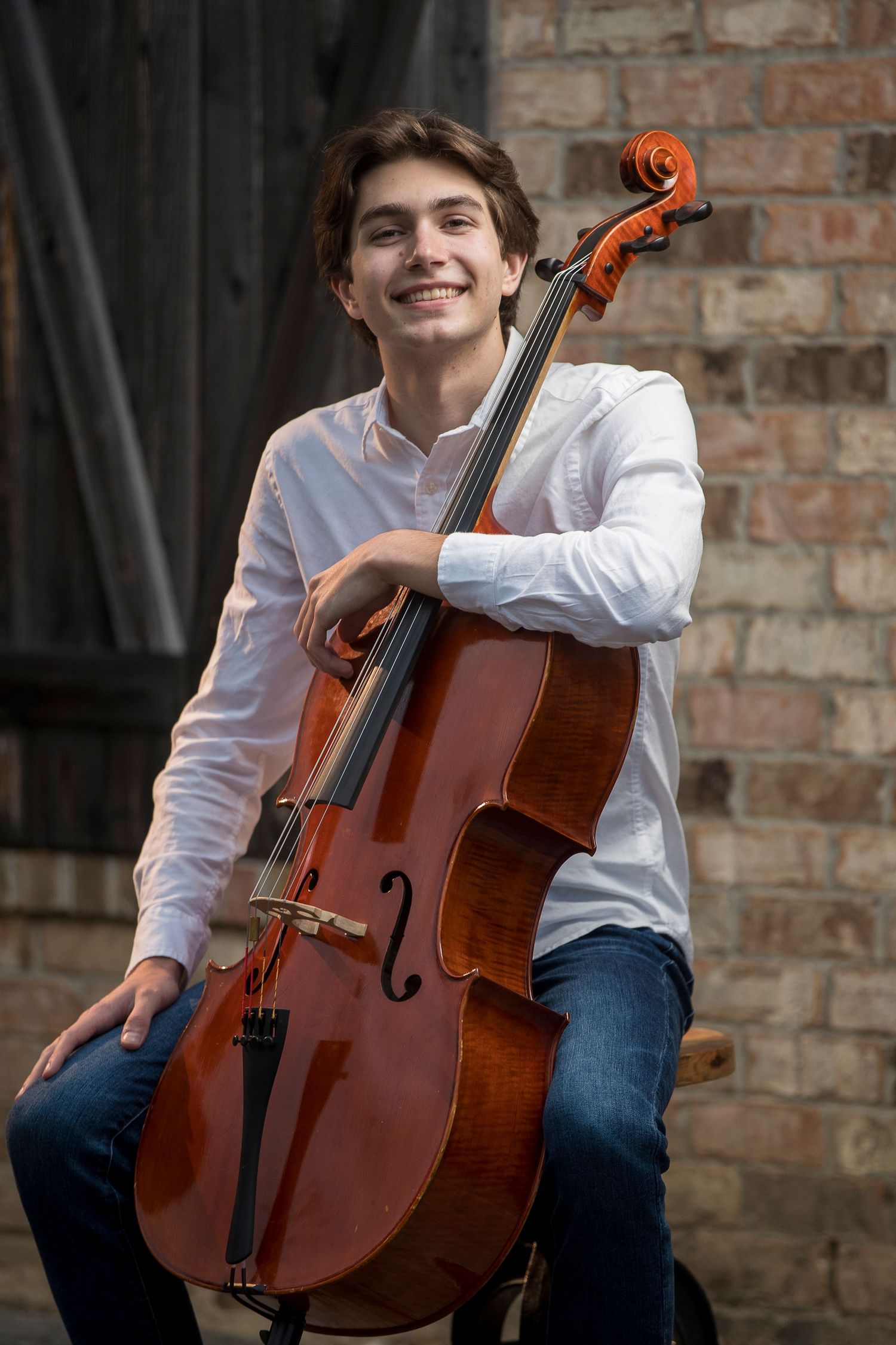 Musician poses with cello against brick wall backdrop in formal attire.