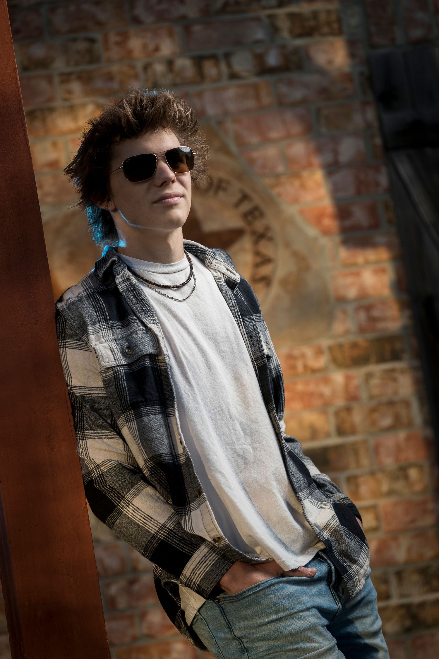 Person in plaid shirt and sunglasses posing against brick wall backdrop.