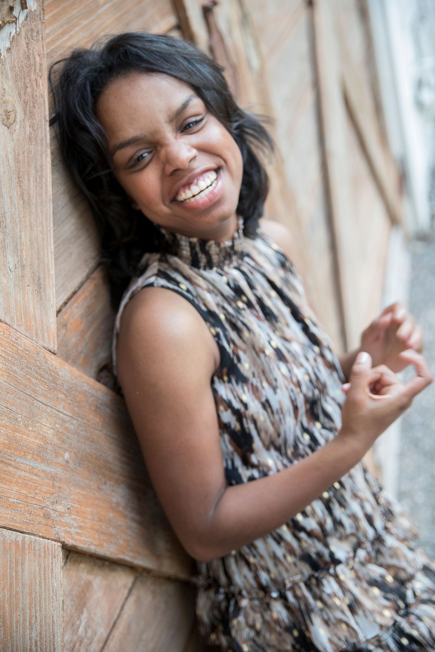 A person in a patterned dress poses joyfully against a rustic wooden wall.