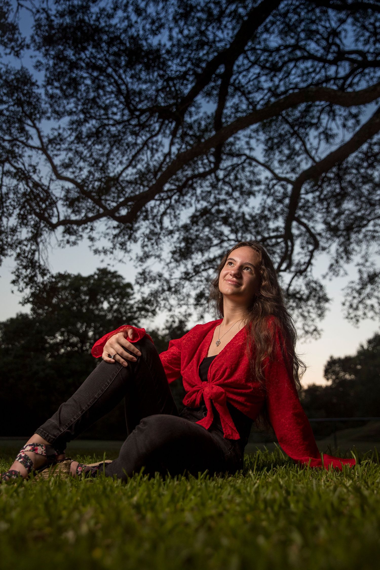 Person in a red dress lying on grass at sunset under tree branches.