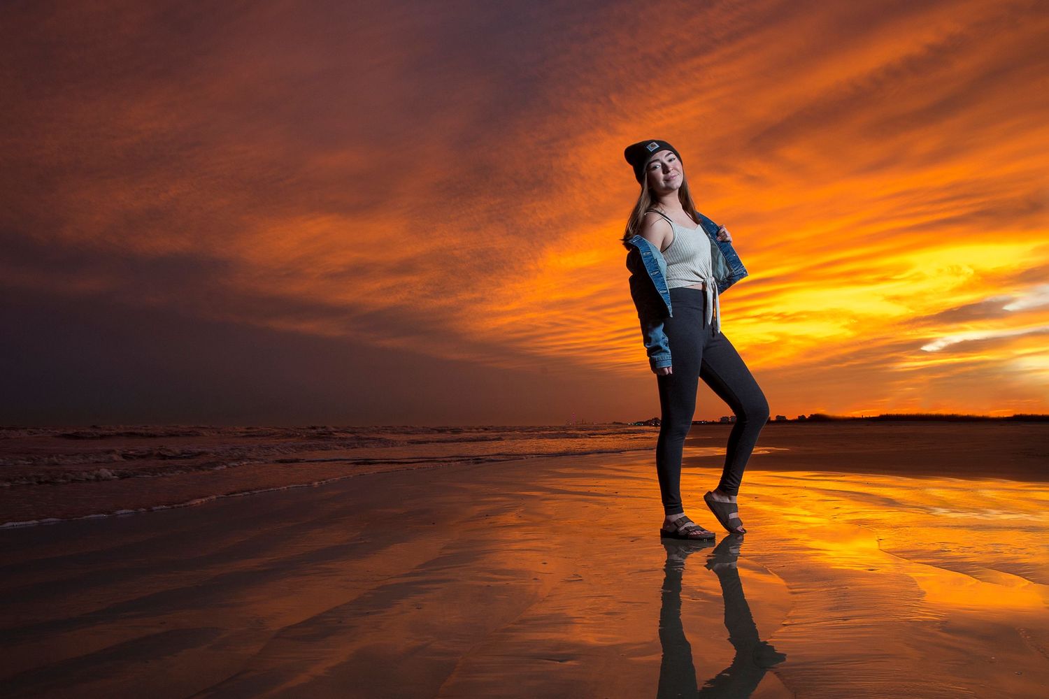 Silhouette of person standing on reflective beach surface during dramatic orange sunset.