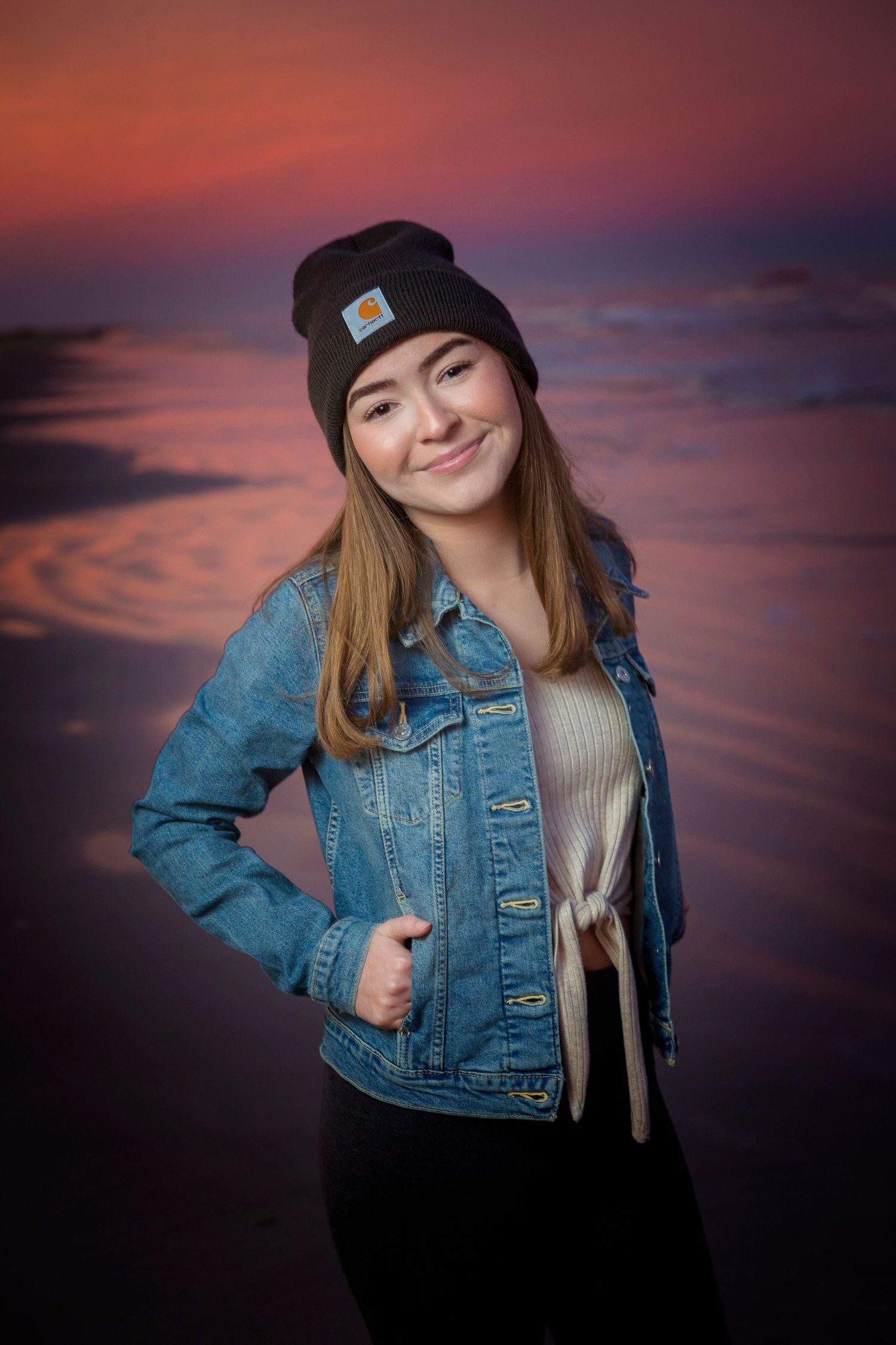 Sunset portrait session wearing denim jacket and beanie against dramatic pink sky.