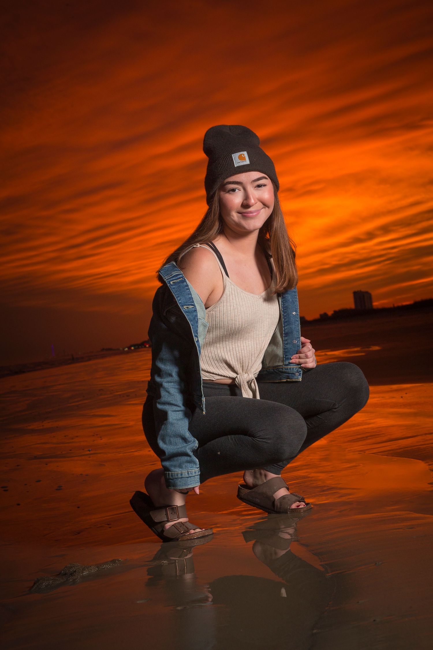Person in beanie and denim jacket crouches on beach during vibrant sunset.