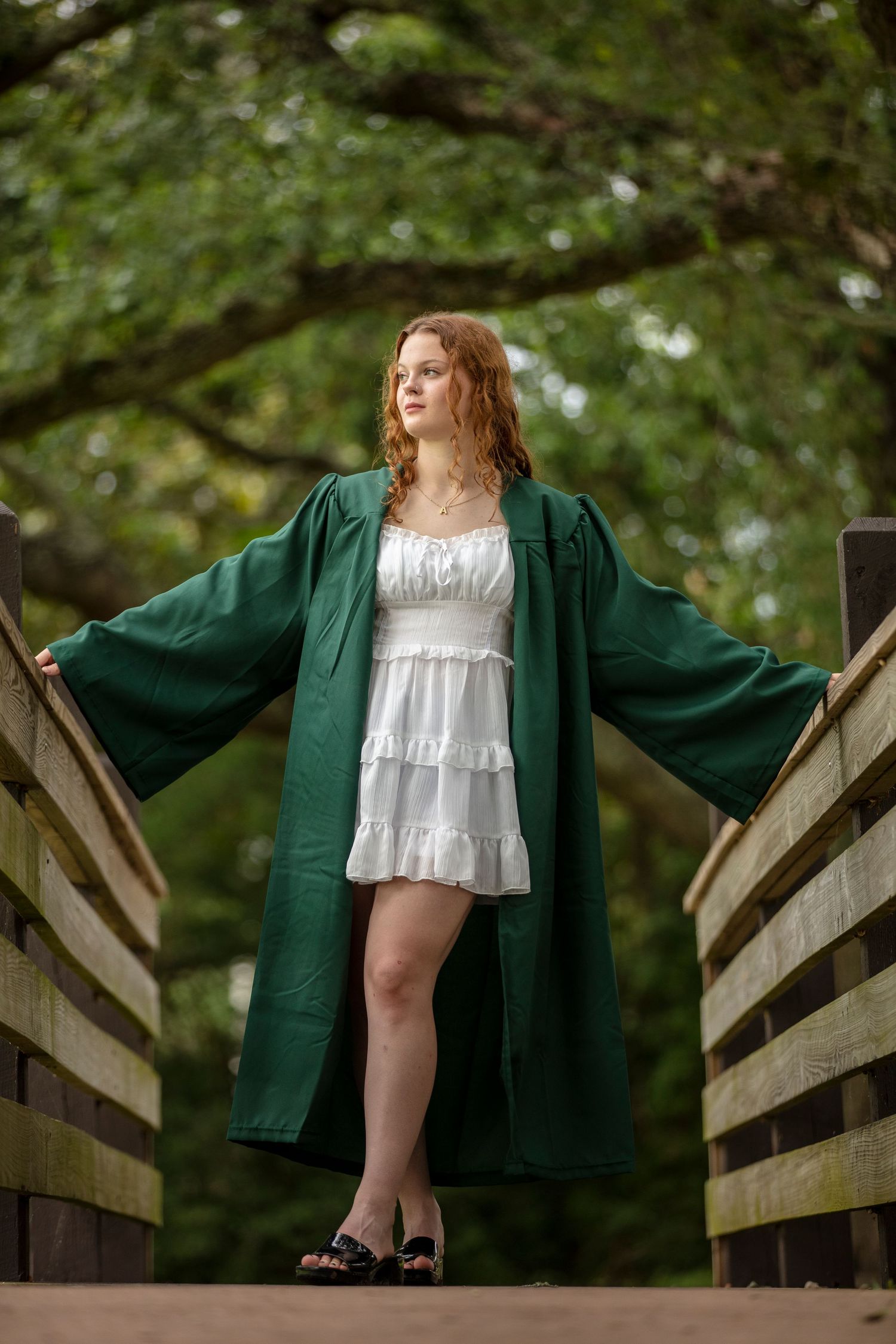 Figure in a white dress and dark green coat poses on a wooden bridge surrounded by lush greenery.